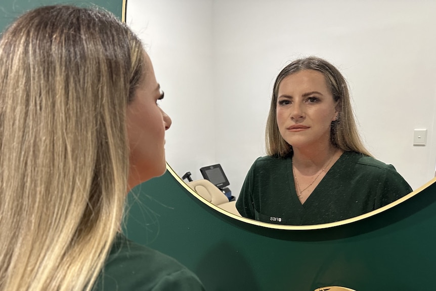 A blonde woman in scrubs looks at herself in a mirror.