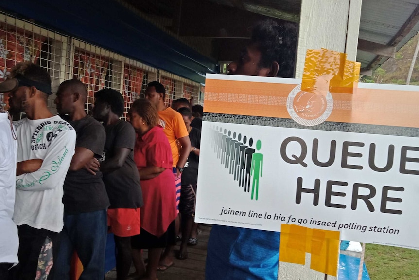 A line of voters wait next a to a sign directing people where to vote.