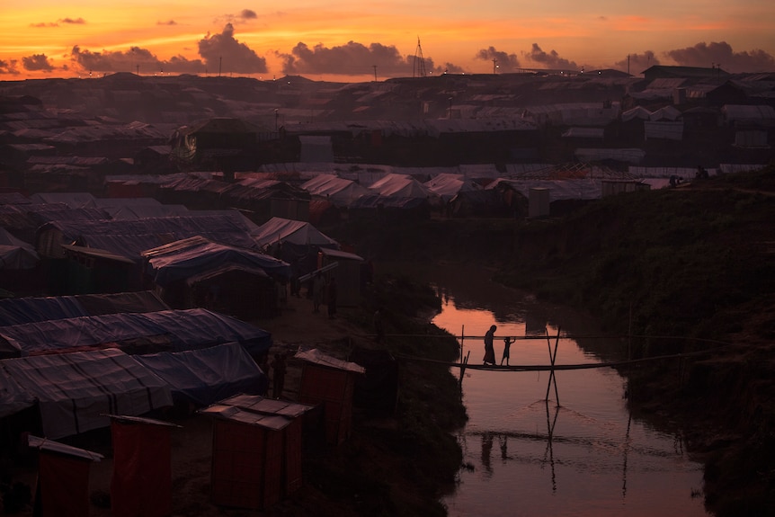 A sunset over a sprawling refugee camp, with two people crossing a bridge over a river