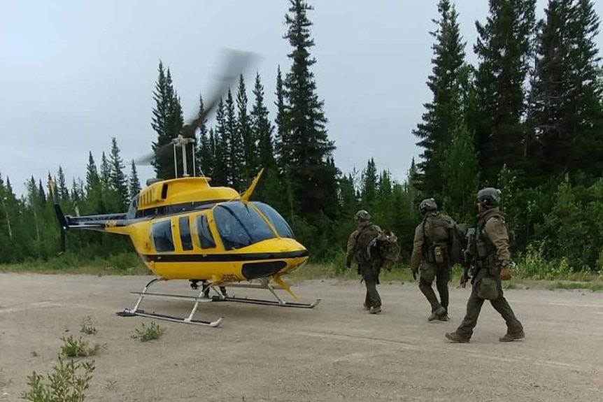 Canadian police officers walk towards a yellow helicopter on a forest road.
