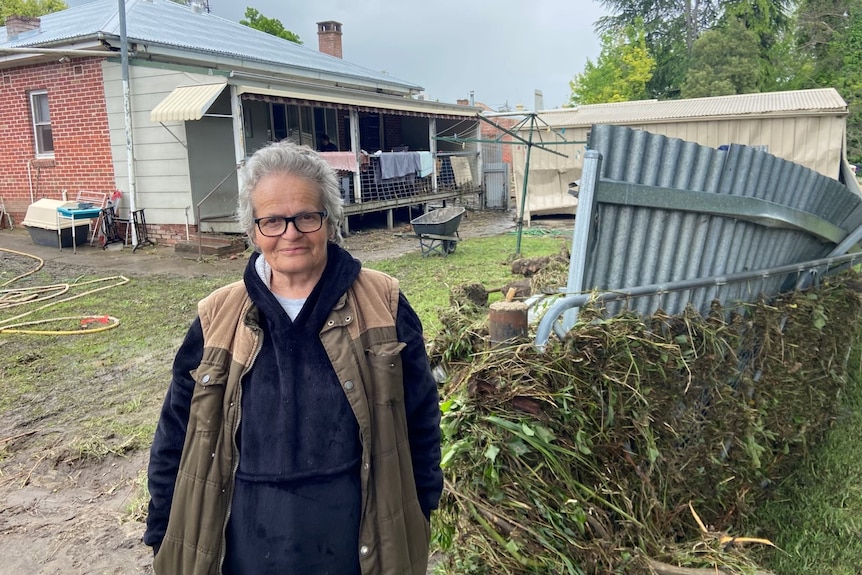 woman with grey hair wearing a jacket stand in front of a flood damaged backyard