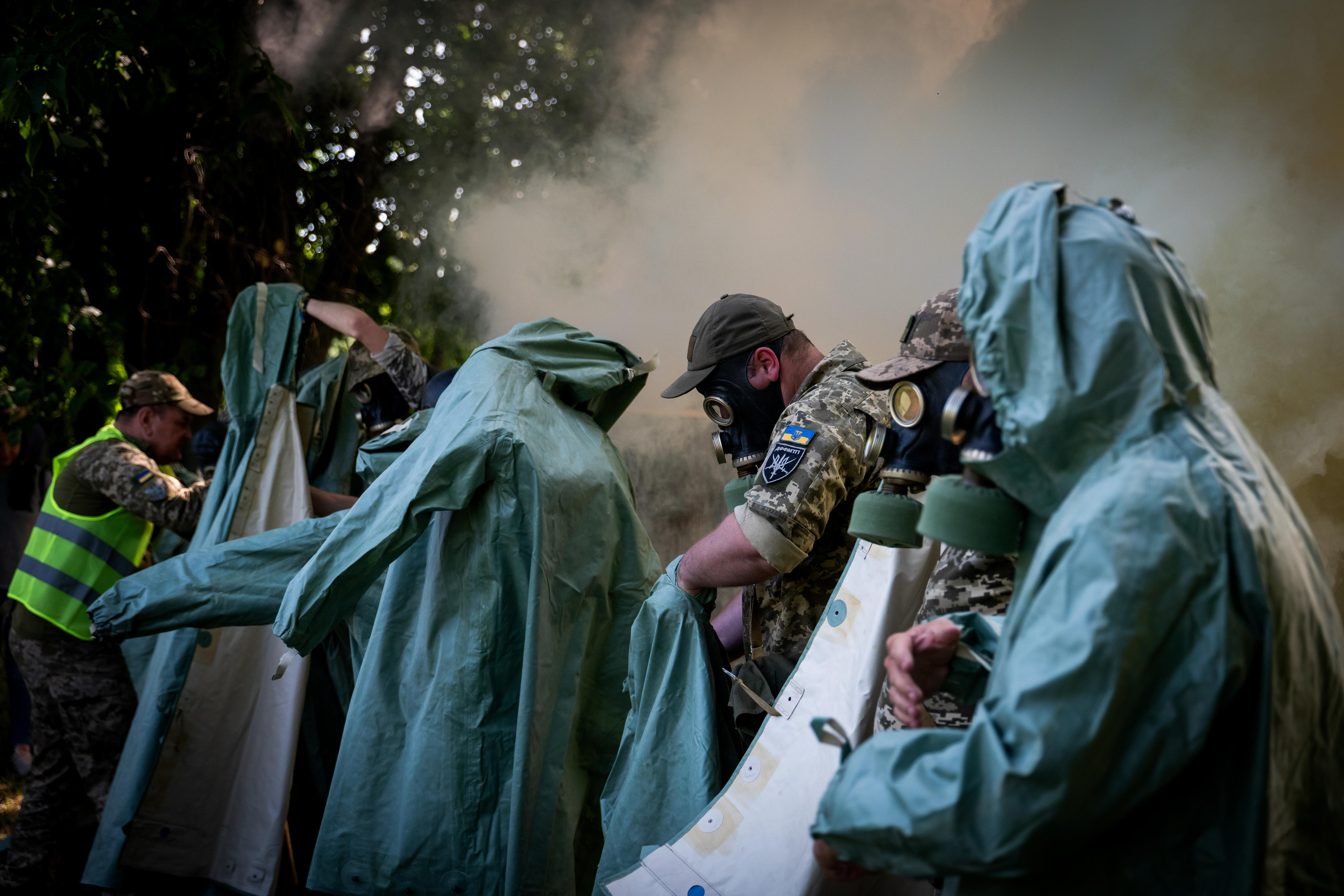 People, some in army fatigues, don turquoise protective suits and gas masks while smoke billows behind them