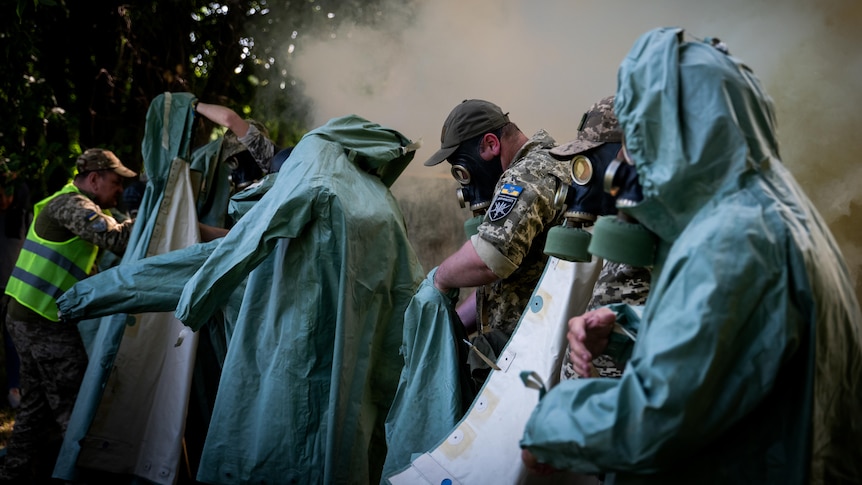 People, some in army fatigues, don turquoise protective suits and gas masks while smoke billows behind them