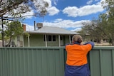 Woman in high-vis shirt leans on backyard fence overlooking rundown weatherboard house, regional Victoria.