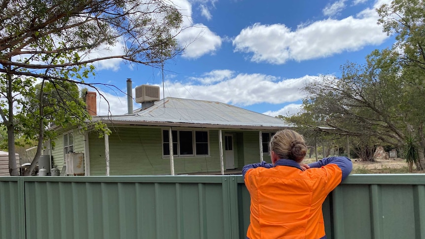 Woman in high-vis shirt leans on backyard fence overlooking rundown weatherboard house, regional Victoria.