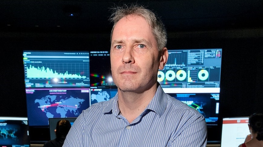 A man wearing a blue business shirt stands with his arms crossed in a dark room filled with computers .