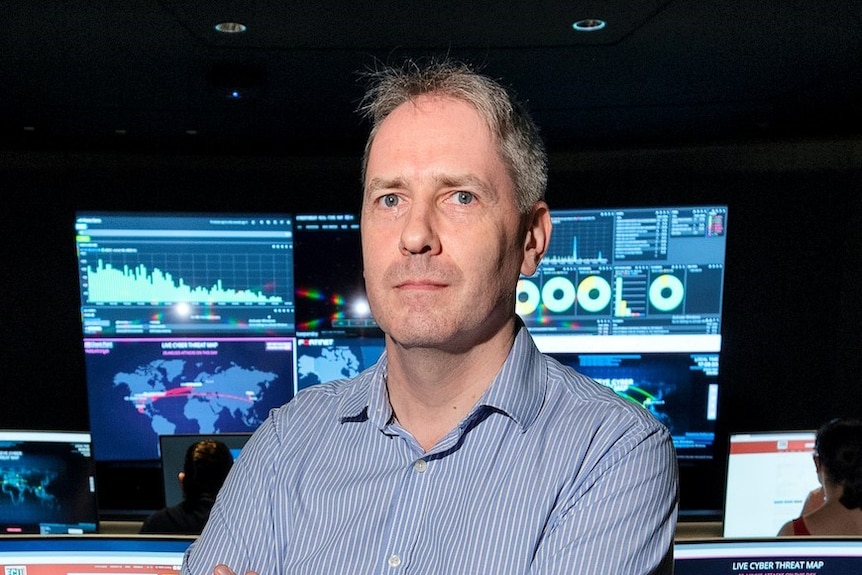 A man wearing a blue business shirt stands with his arms crossed in a dark room filled with computers .