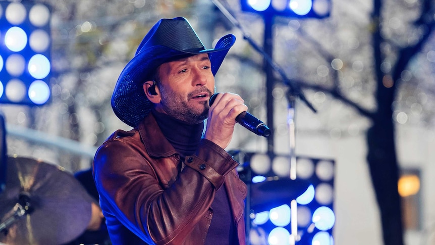 Tim McGraw in a black country style had and lwather jacket sings into a handheld microphone in front of a drummer