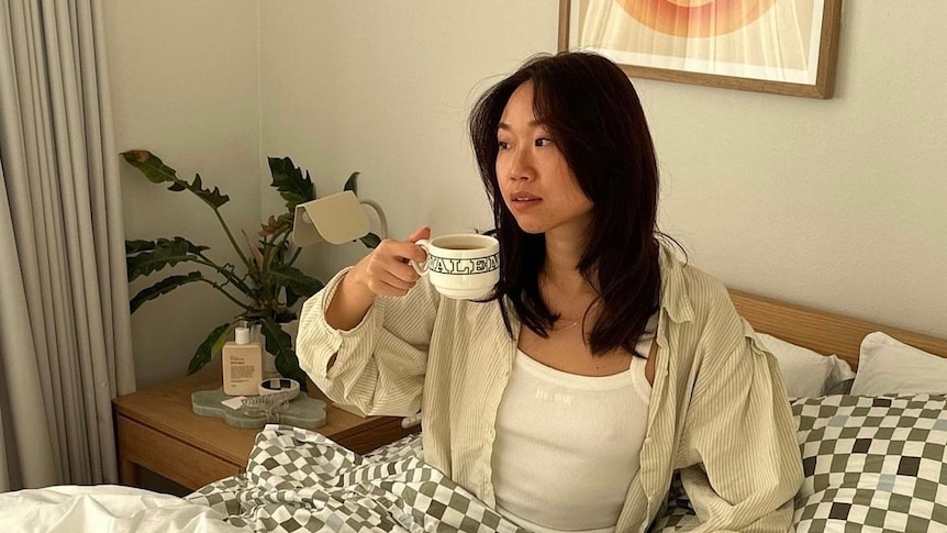 A young woman with dark hair sits up in bed, ceramic coffee cup in hand.