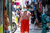 A woman in a red sarong and face mask walks through a narrow street