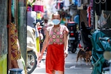 A woman in a red sarong and face mask walks through a narrow street
