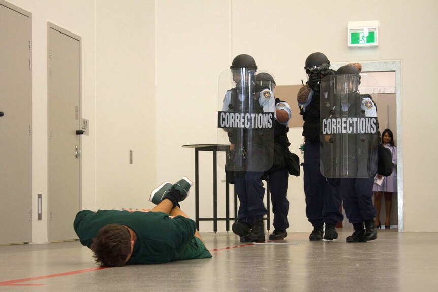 Prison officers holding shields and wearing protective gear approach a prisoner lying flat on the ground.