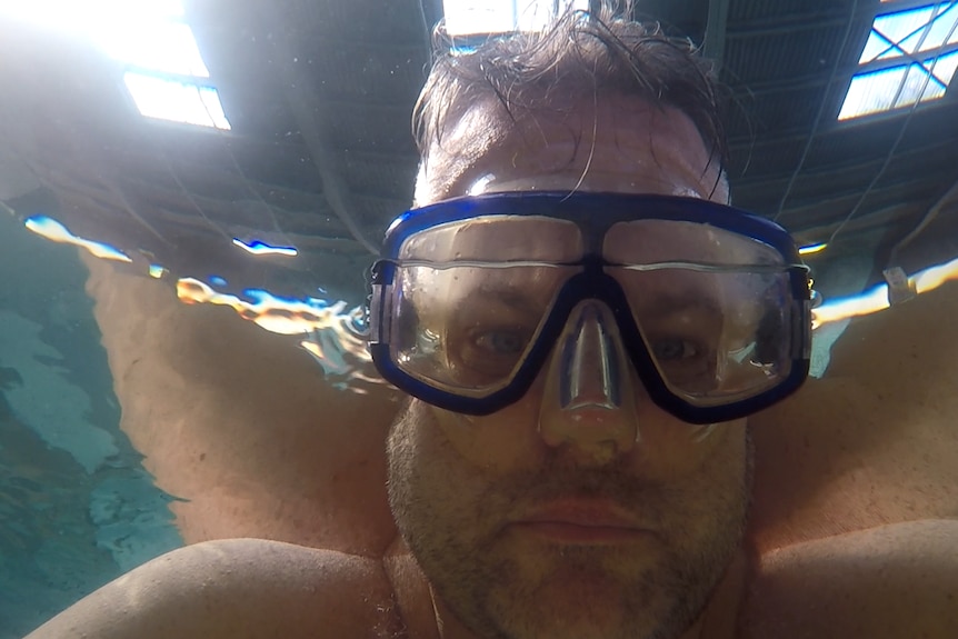 A reporter wearing blue goggles trying to hold his breath underwater.