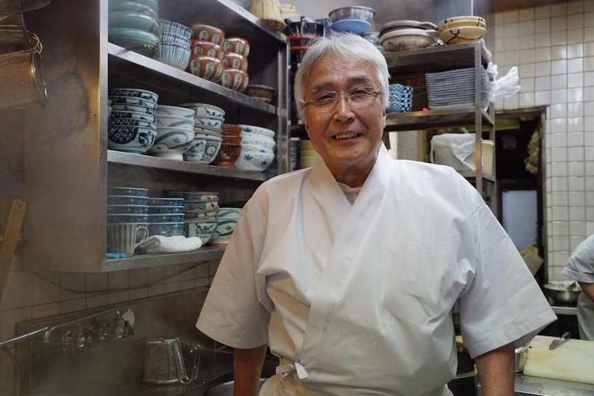 Hideki Tanaka stands in his restaurant kitchen smiling in front of bowls.