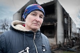 A woman in a puffer coat with a bright blue scarf tied around her head stands in front of a burned out building 