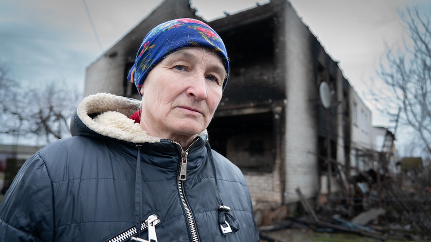 A woman in a puffer coat with a bright blue scarf tied around her head stands in front of a burned out building 