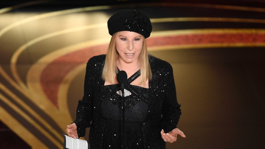 Barbra Streisand stands on stage and speaks into a microphone to present an award