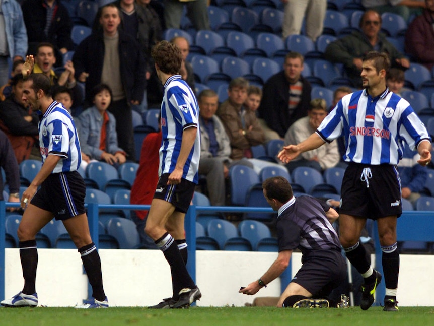 A Premier League footballer (far left) walks away after being sent off, as teammates watch and the referee sits on the ground.