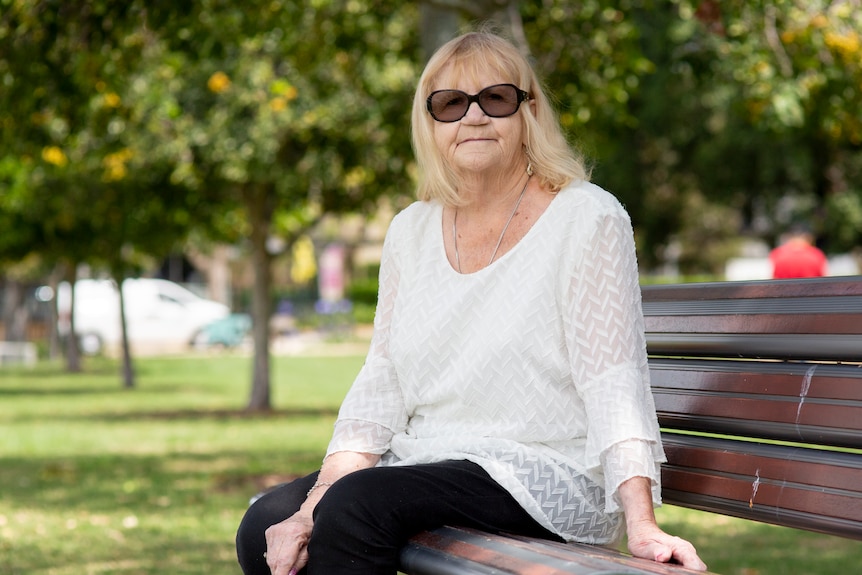 An older woman with blonde hair and wearing sunglasses and a white shirt sits outside on a park bench.