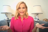Reese Witherspoon in a pink shirt, sitting on her couch and shrugging