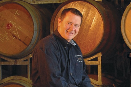 A dark-haired man, smiling, standing in a wine cellar.