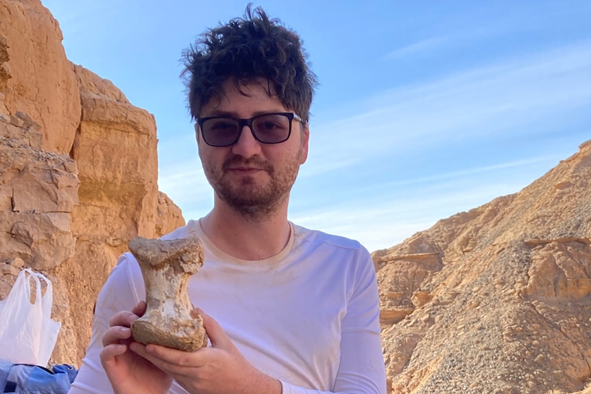 A young man with a mop of brown hair, light sunglasses, long-sleeved white t-shirt, holds a dinosaur bone.