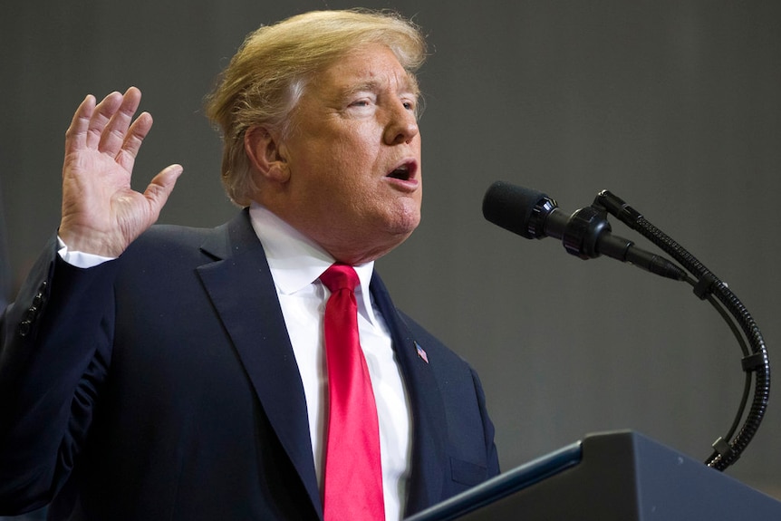 A man with orange-brown hair wearing a navy suit and red tie raises his right hand as he speaks into a microphone