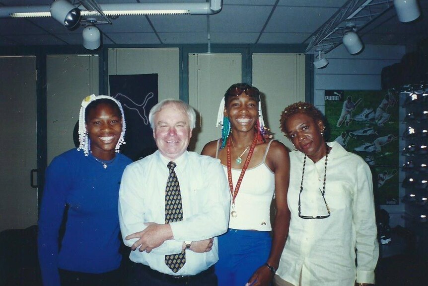 An older man stands smiling with Serena and Venus Williams and their mother.