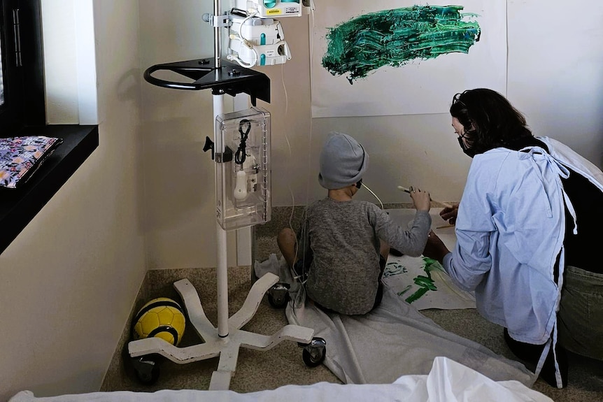 A young boy attached to drips, sits on the floor of his hospital room with a teacher, doing a painting.