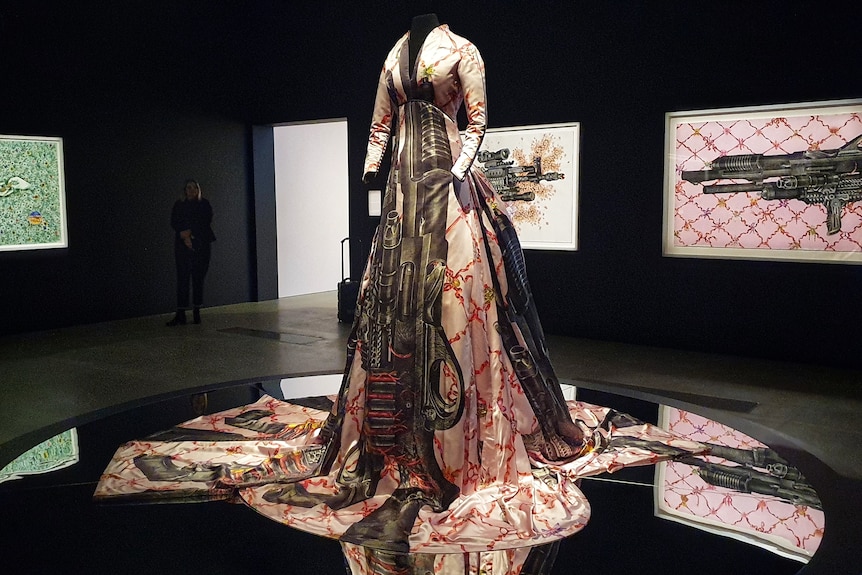 A gown printed with images of guns displayed in a dark exhibition room.