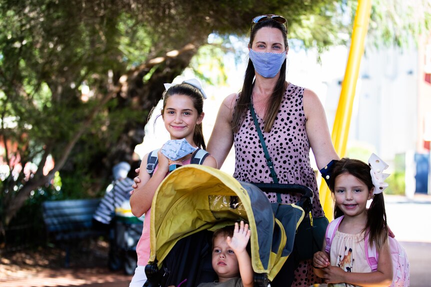 A woman with a mask on stands next to her two daughters and a pram with her son in it.