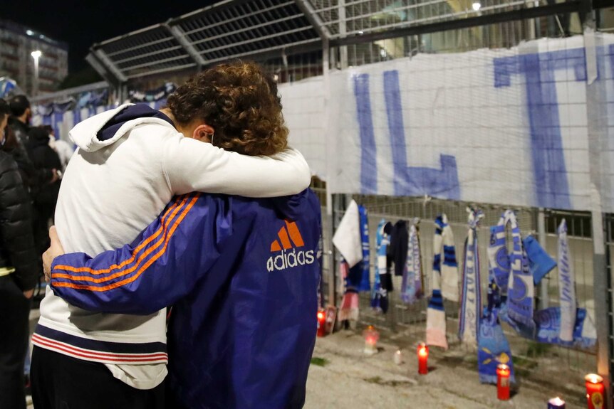 Two people hug as one rest their head on the other's shoulder near candles and scarves laid in tribute to Diego Maradona