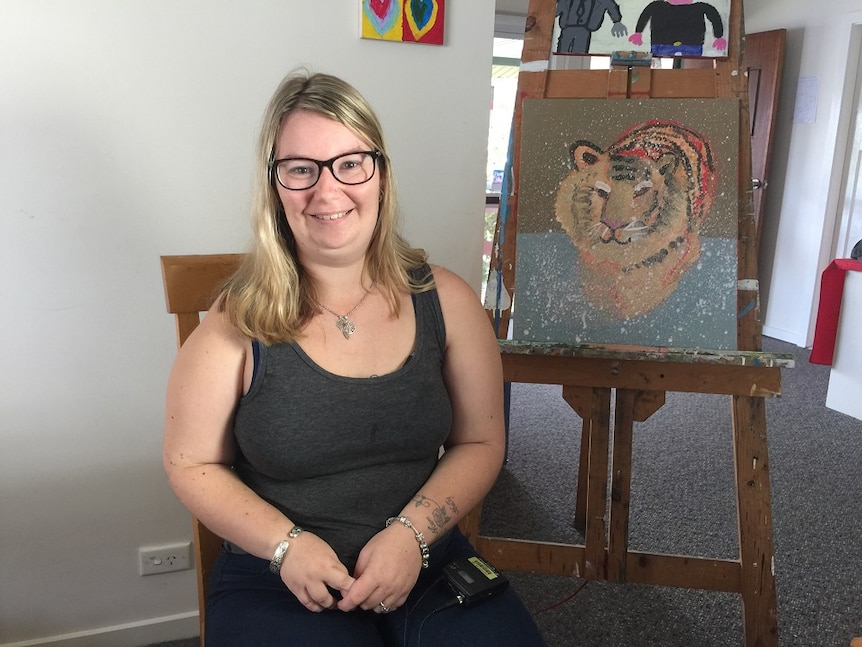 A young woman with blonde hair and glasses standing in front of an easel.