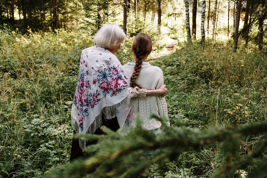 White-haired older woman hugging a young teenage girl standing in a green forest.
