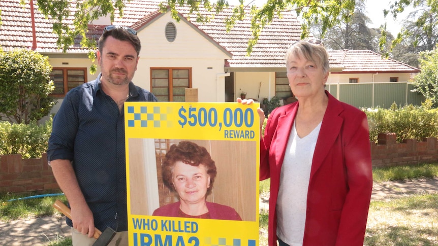 John and Liz stand holding a sign promising a $500,000 reward for helping identify the killers.