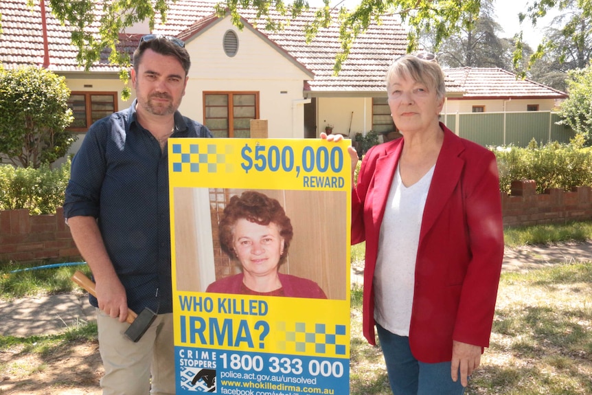 John and Liz stand holding a sign promising a $500,000 reward for helping identify the killers.