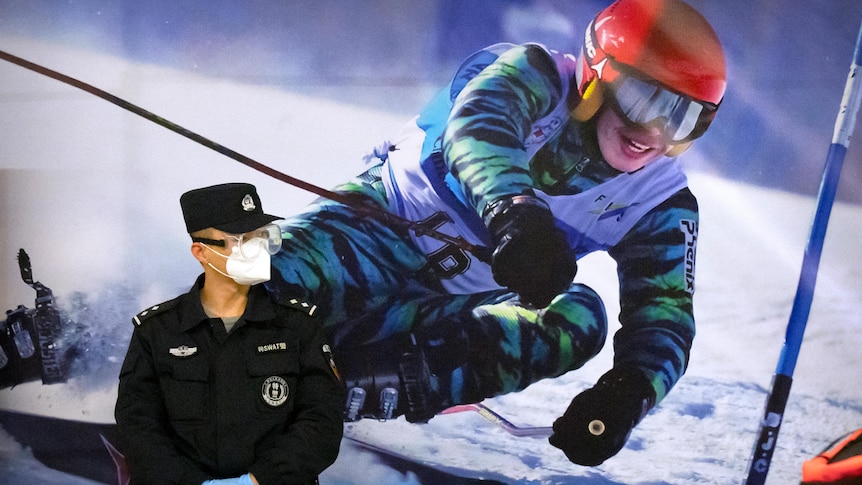 A police officer wearing a face mask stands next to a poster of a skier