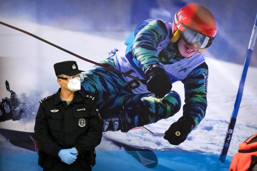 A police officer wearing a face mask stands next to a poster of a skier
