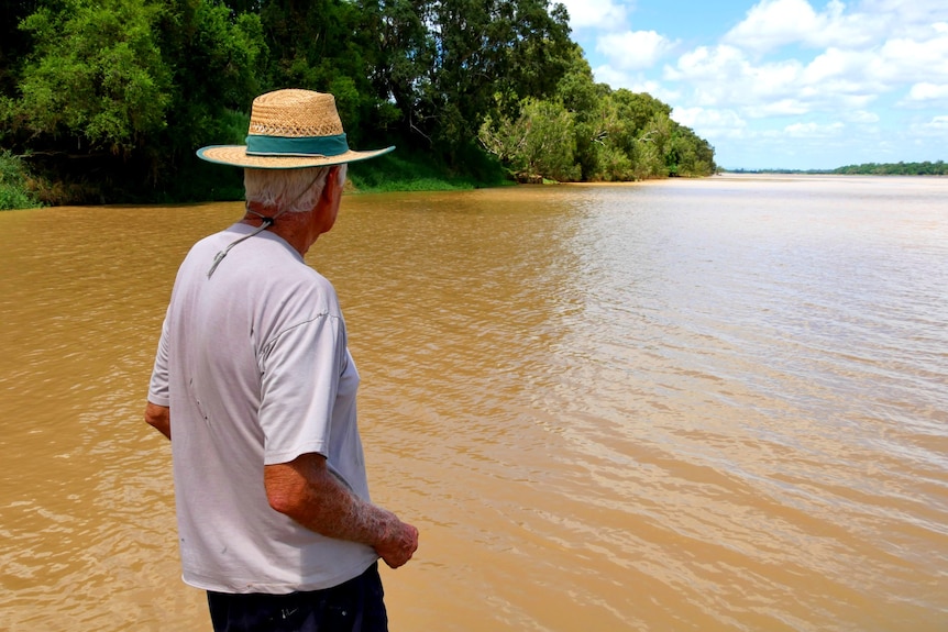 A farmer with a brown hat looks out at brown water of the Burdekin River. 