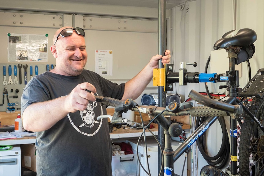 A man smiles while holding a bike that is hanging from a metal bar in a workshop.