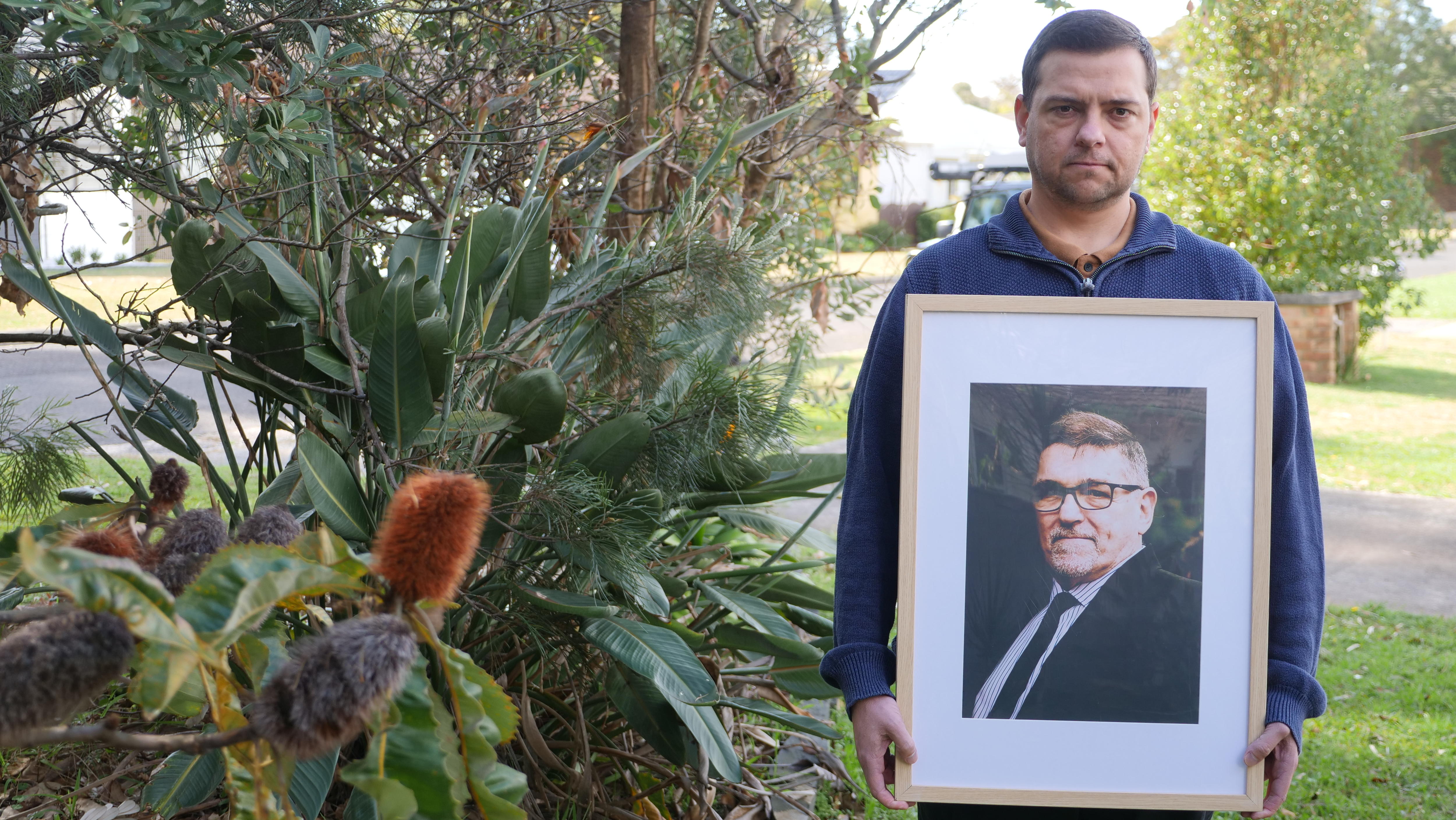 image of middle aged man holding a large photo of an older man with a stern expression in a garden