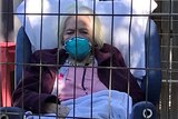 An elderly woman with a mask on behind a metal fence.