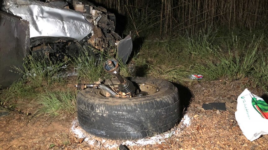 A car wheel ripped off the chassis, with the front of the wrecked car in the background.