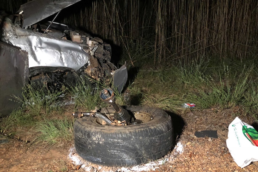 A car wheel ripped off the chassis, with the front of the wrecked car in the background.