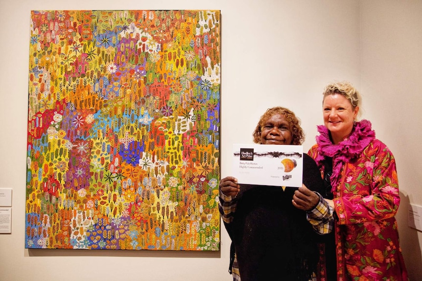 Two women stand next to an Indigenous painting and display the award it received.