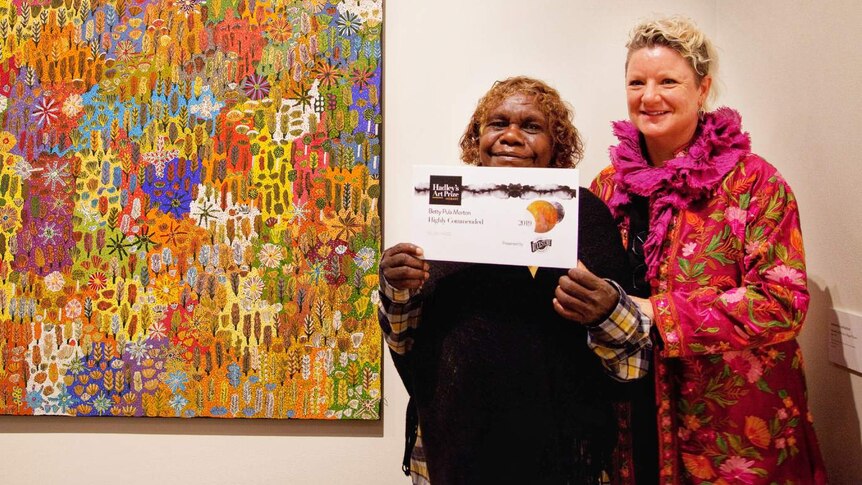 Two women stand next to an Indigenous painting and display the award it received.