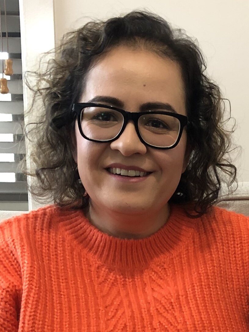 A photo of a woman with curly brown hair and glasses smiling at the camera while wearing an orange knit jumper.
