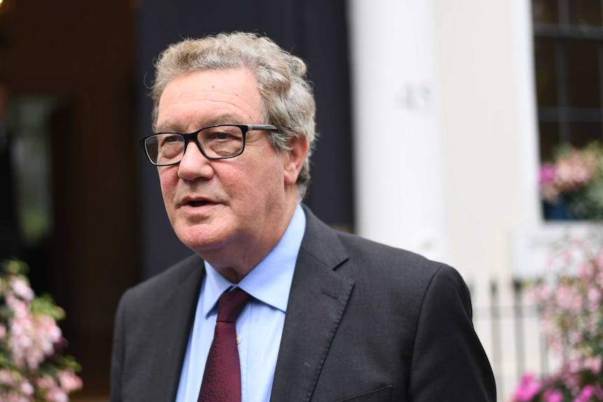 Mr Downer is wearing thick rimmed black glasses, a dark grey suit, blue shirt and maroon tie.