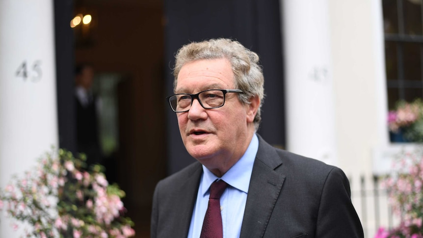 Mr Downer is wearing thick rimmed black glasses, a dark grey suit, blue shirt and maroon tie.