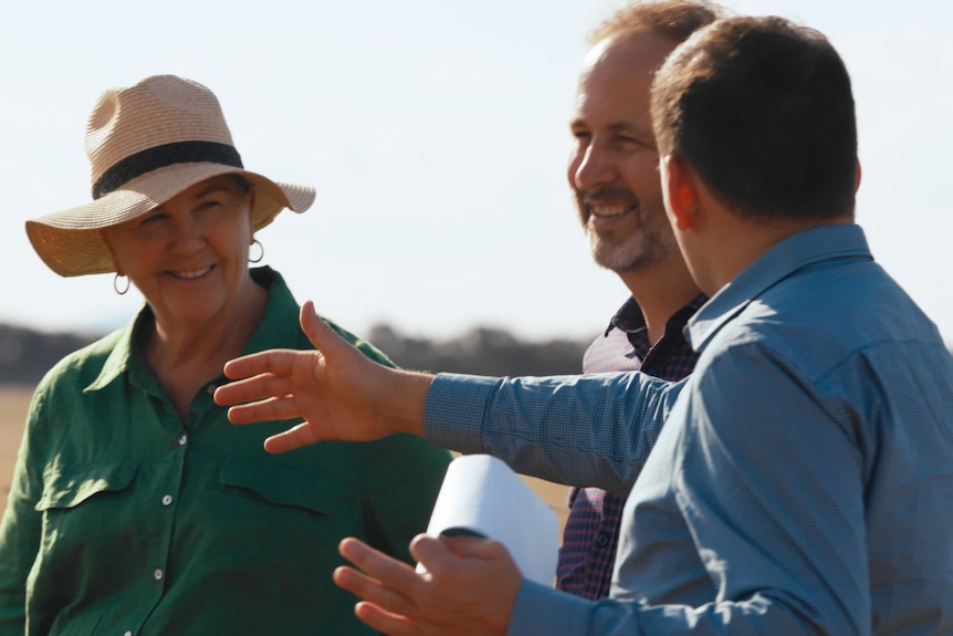 Two men and a woman stand on a farm smiling, and discussing something.